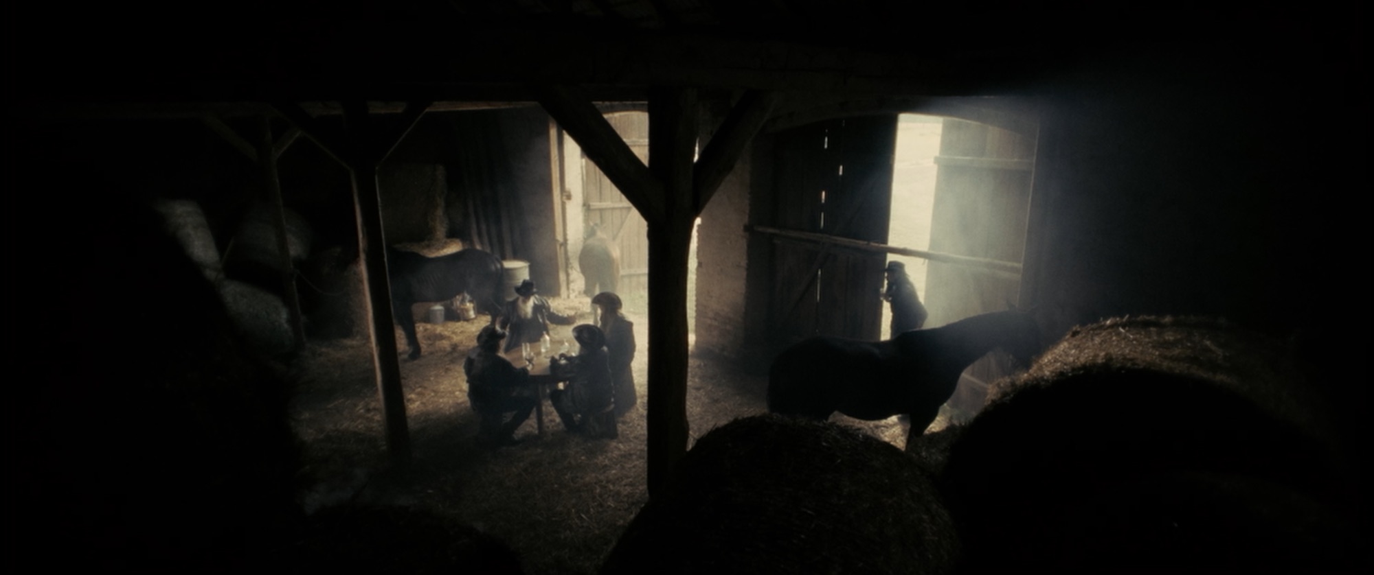 Barn interior seen past hay bales - figures gathered at a table in a shaft of light, horses in the gloom - Amon Amarth